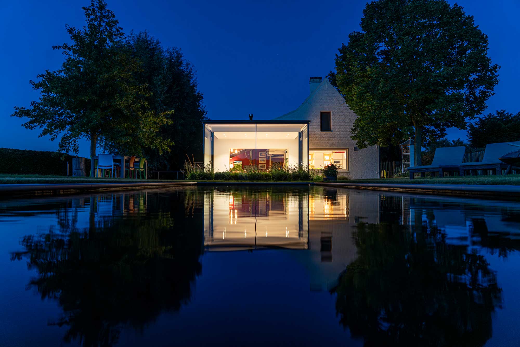  Nachtaufnahme eines modernen, beleuchteten Wintergarten-Anbaus in Mülheim, der sich im ruhigen Wasser eines Pools spiegelt. 