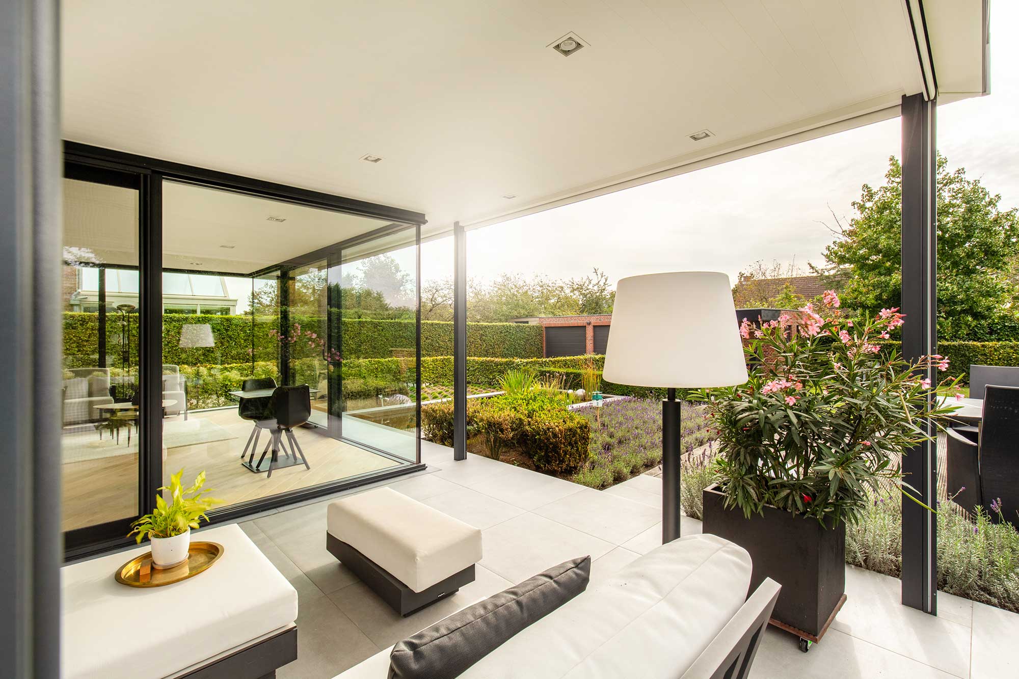  Moderne Terrasse in Mülheim mit Wintergarten-Anbau, weißer Lounge, Nicholls Brecon Tischleuchte und Blick in den gepflegten Garten. 