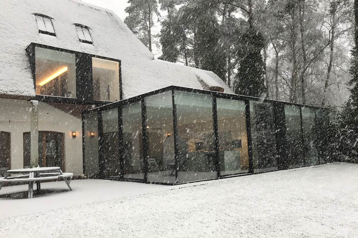 Winterlicher Glas-Wintergarten in Mülheim mit schwarzem Rahmen an einem schneebedeckten Haus mit Reetdach. Verschneiter Garten mit Bäumen.