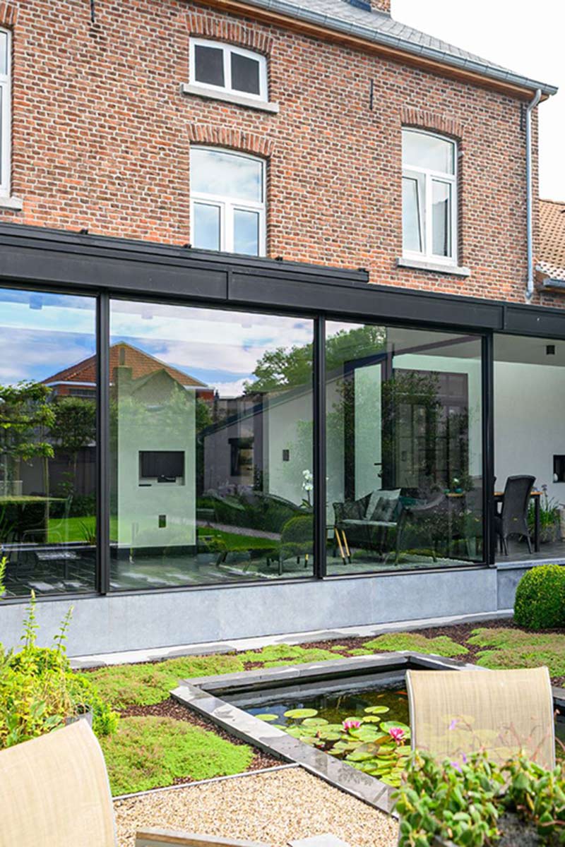 Moderner Glas-Wintergarten in Mülheim an einem Backsteinhaus mit Blick in einen grünen Garten mit Teich und stilvollen Gartenmöbeln.