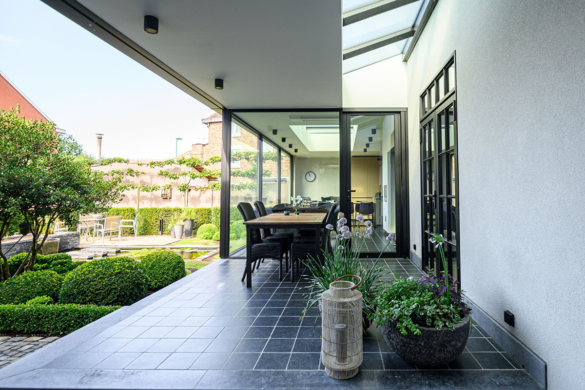 Moderner Wintergarten in Mülheim mit schwarzem Rahmen, Esstisch auf dunklen Fliesen und Blick in einen formalen Garten mit Wasserbecken.