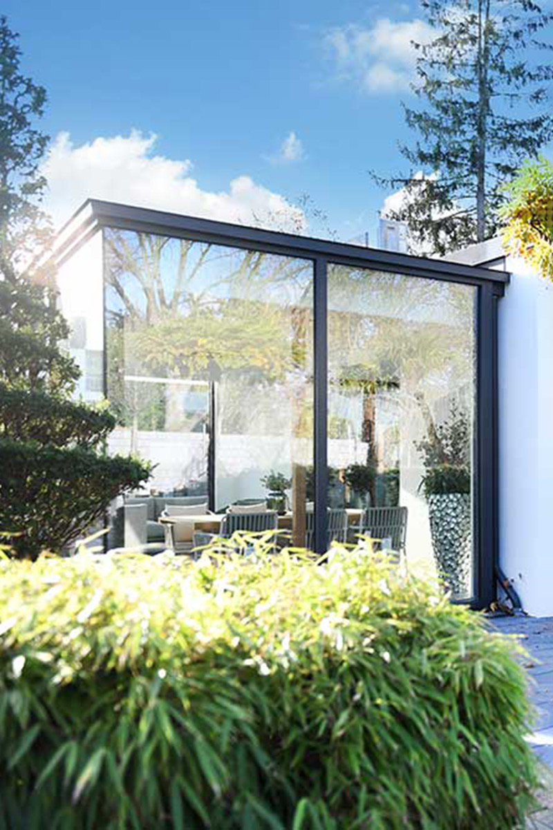 Moderner Glas-Wintergarten in Mülheim mit schwarzem Rahmen, umgeben von üppigem Bambus und einem stilvollen, kleinen Baum unter blauem Himmel.
