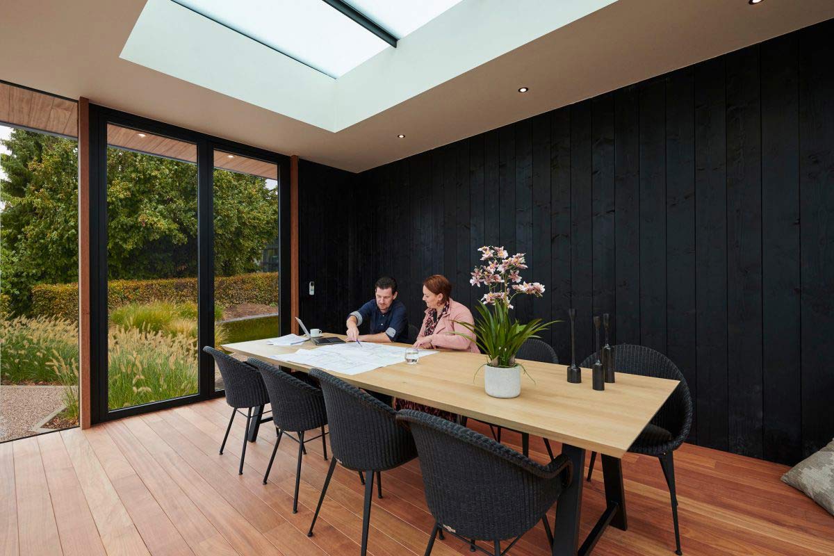 Heller Arbeitsbereich in einem modernen Gartenpavillon in Mülheim mit Holzboden, schwarzer Holzwand, großem Tisch, zwei Personen und Blick in den Garten.