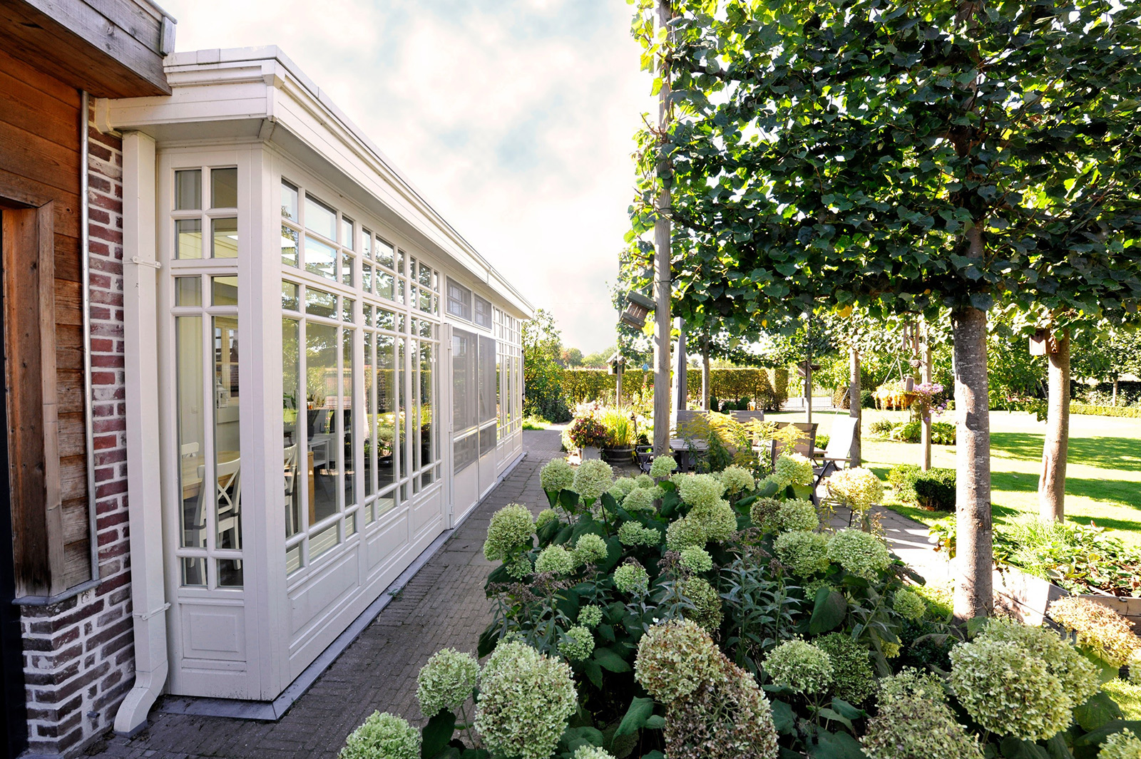 Heller, weißer Wintergarten an einem Backsteinhaus mit gepflastertem Weg und blühenden Hortensien im Garten.