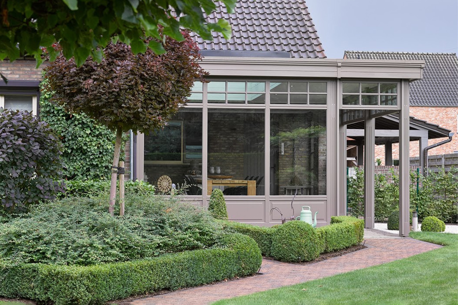 Elegante, graue Terrassenüberdachung/Veranda mit Glasfront und Sprossenfenstern, umgeben von einem gepflegten Garten mit Buchsbaumhecken.